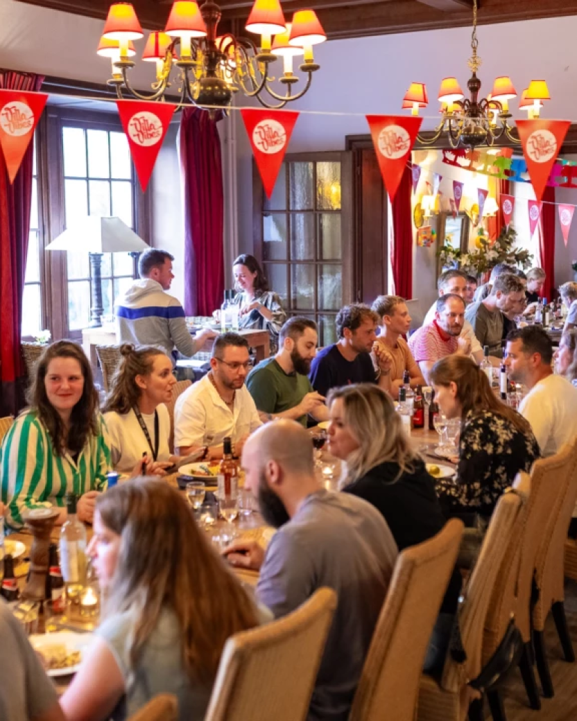 Groep reisgenoten eet samen aan lange tafels in een sfeervolle gezamenlijke ruimte.