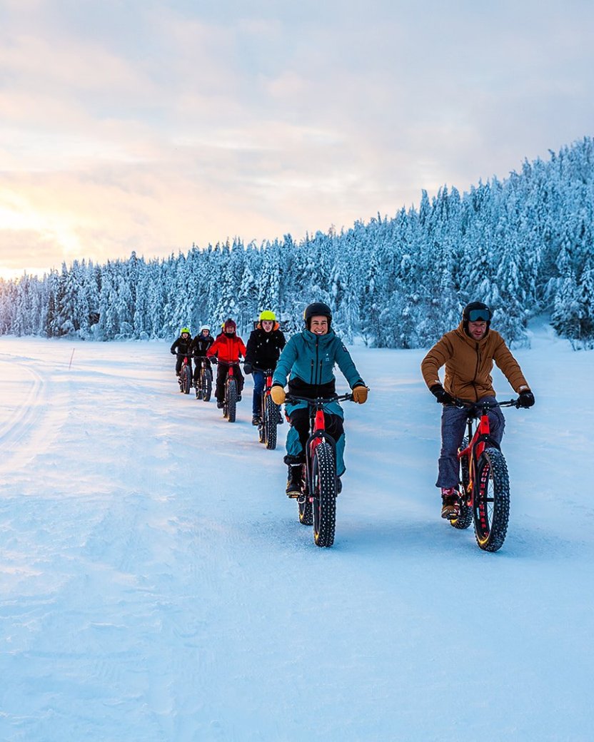 Groep reisgenoten fietst met fatbikes door een besneeuwd landschap bij zonsondergang.