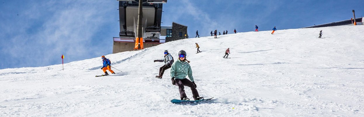 Snowboarder daalt af over een zonnige skipiste met stoeltjesliften op de achtergrond.