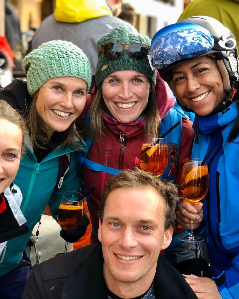 Groep wintersporters poseert lachend met drankjes tijdens de après-ski.