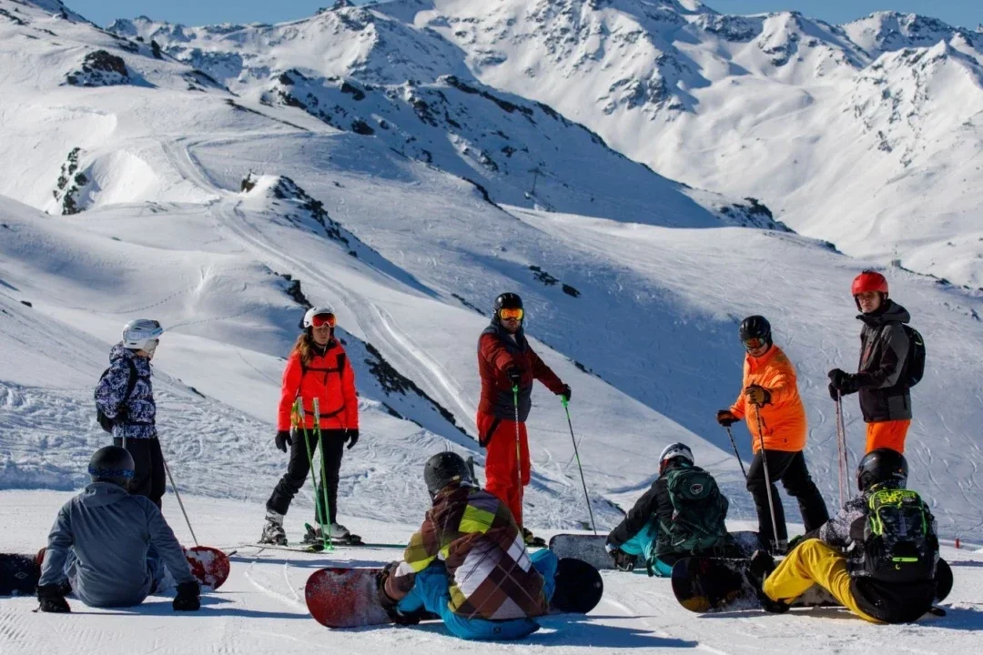 Groep skiërs en snowboarders pauzeert samen op de piste met besneeuwde bergen op de achtergrond.