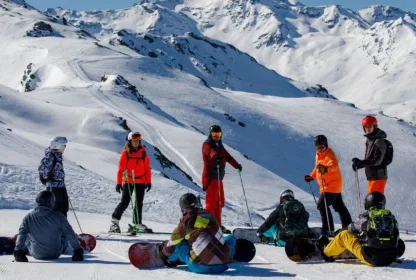 Groep skiërs en snowboarders pauzeert samen op de piste met besneeuwde bergen op de achtergrond.