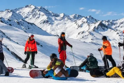 Groep skiërs en snowboarders pauzeert samen op de piste met besneeuwde bergen op de achtergrond.