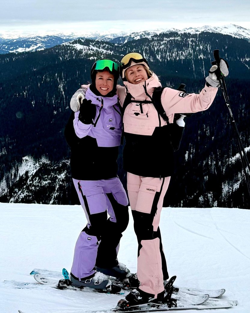 Twee skiërs poseren samen op de piste met besneeuwde bergen op de achtergrond.