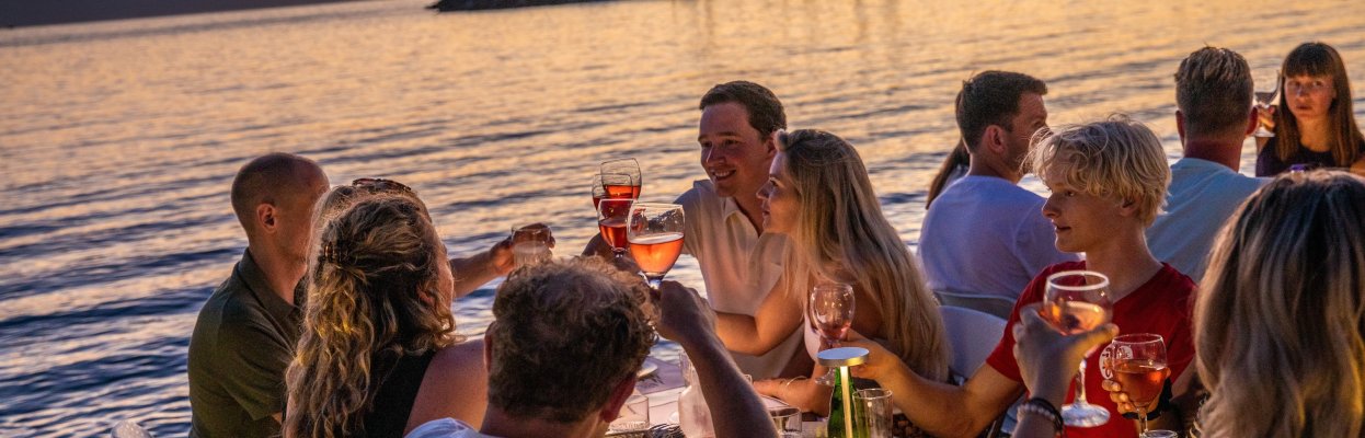 Reisgenoten proosten met elkaar tijdens een diner aan het water met zonsondergang.