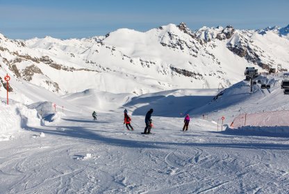 Wintersporters dalen af over een brede piste met besneeuwde bergen op de achtergrond.