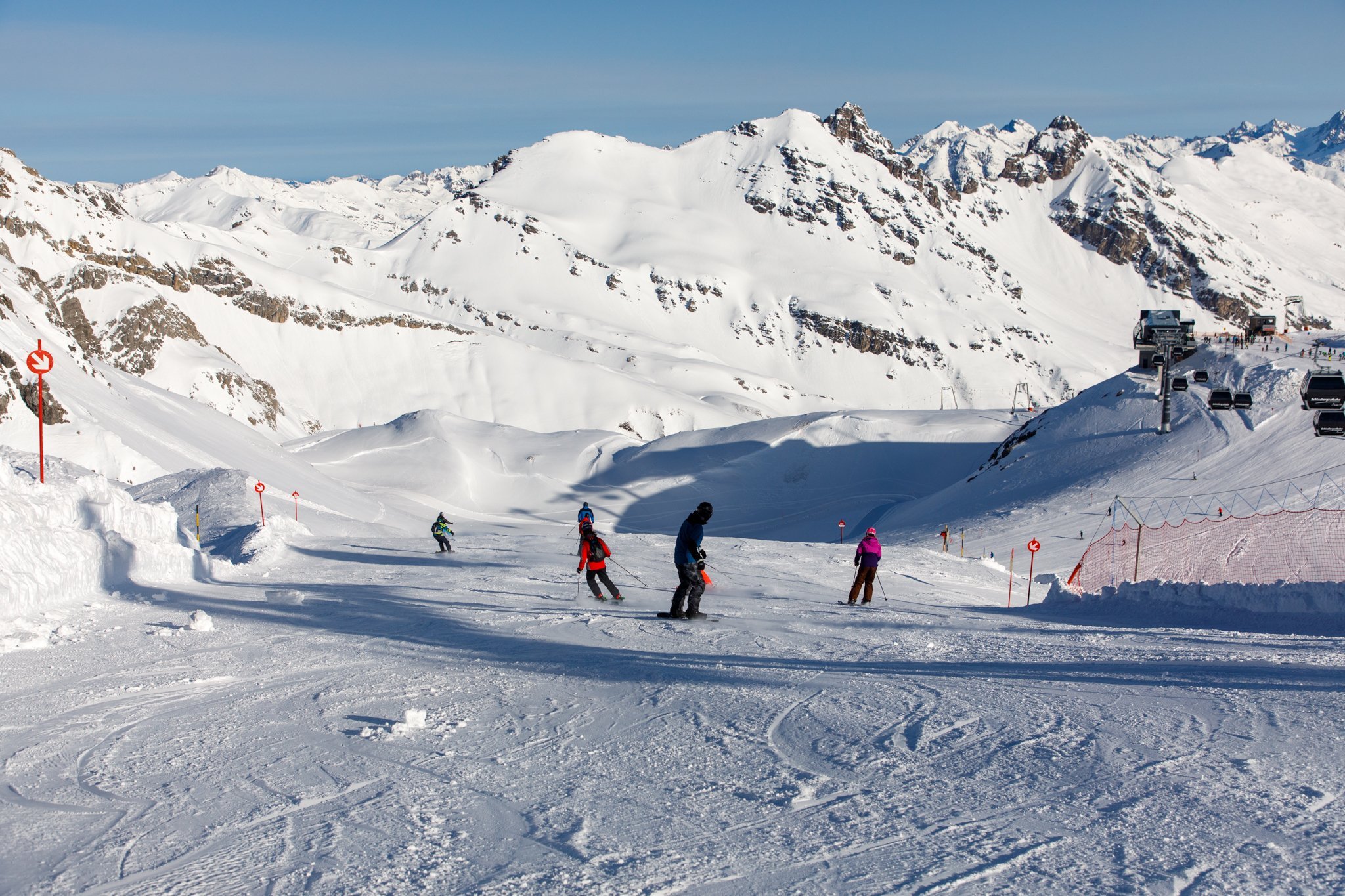 Wintersporters dalen af over een brede piste met besneeuwde bergen op de achtergrond.