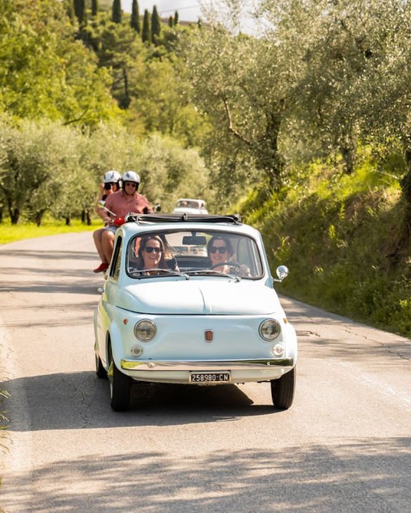 Reisgenoten rijden in een klassieke Fiat door het Toscaanse landschap, met scooters achter hen.