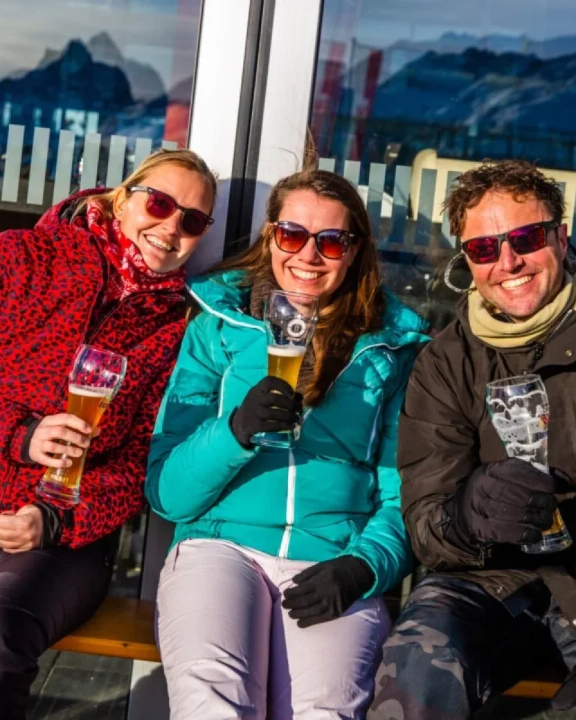 Drie lachende mensen die in de zon op het terras een biertje drinken.