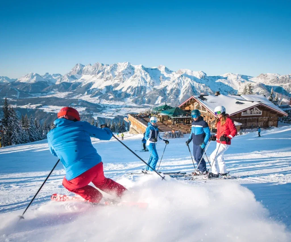 Skiërs op een zonnige piste met een berghut en besneeuwde bergtoppen op de achtergrond.