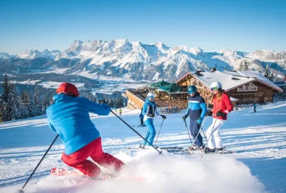 Skiërs op een zonnige piste met een berghut en besneeuwde bergtoppen op de achtergrond.