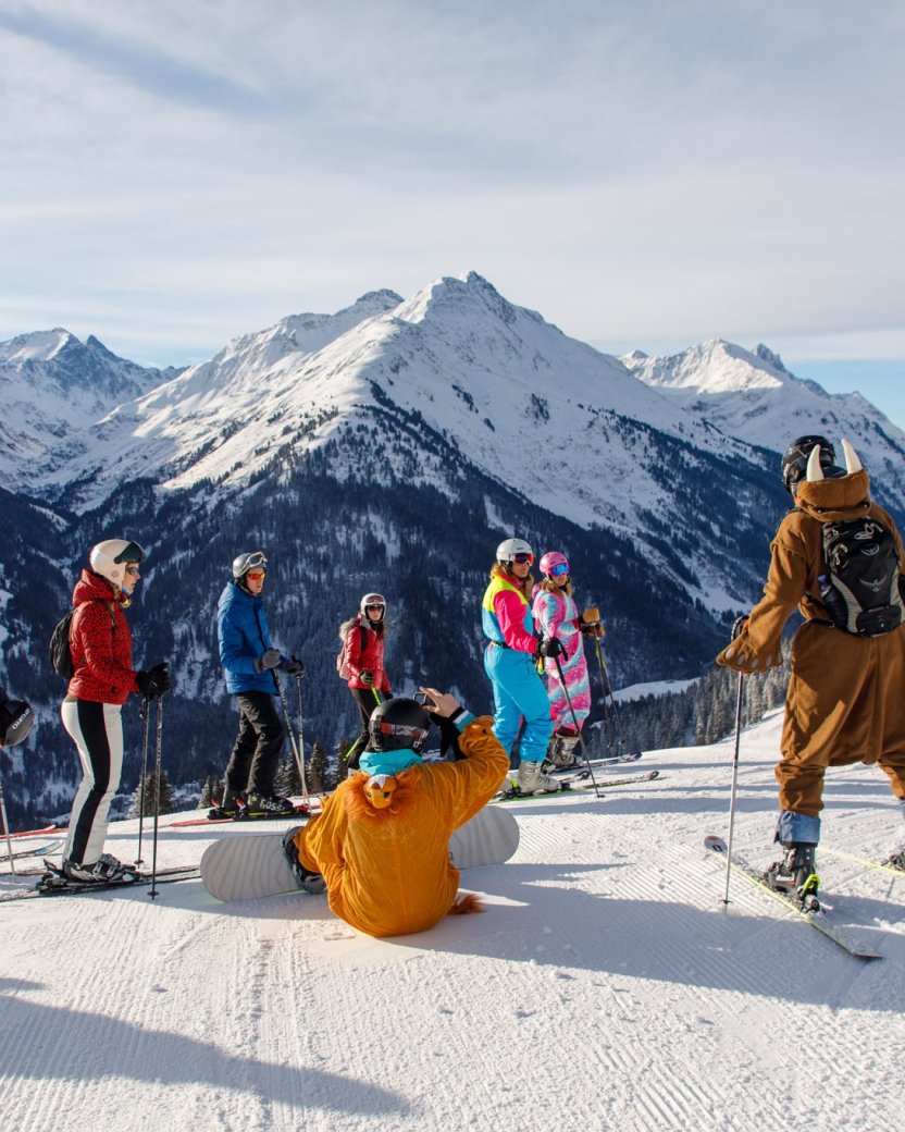 Groep skiërs en snowboarders pauzeert op de piste met uitzicht op de besneeuwde bergen.