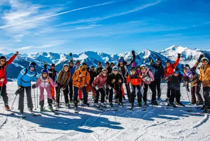 Grote groep wintersporters poseert samen op de piste met besneeuwde bergen op de achtergrond.
