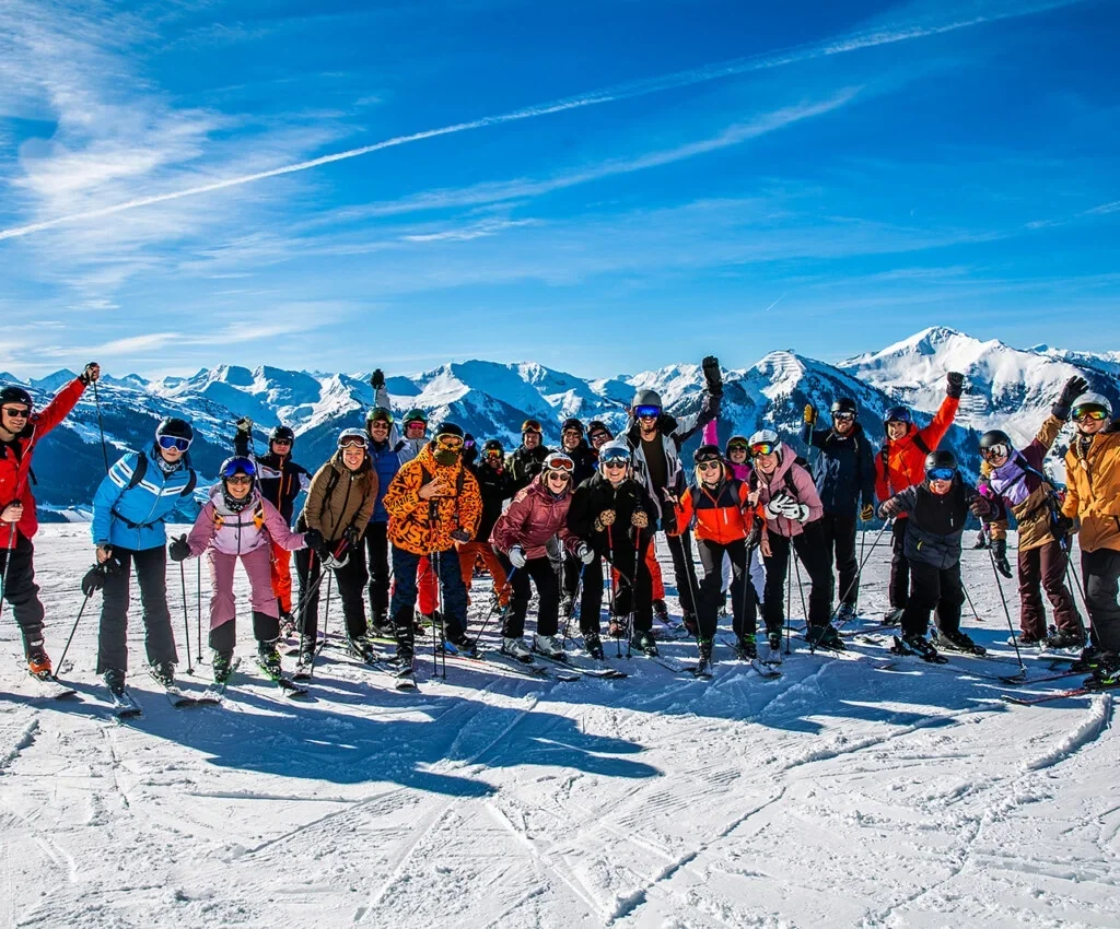 Grote groep wintersporters poseert samen op de piste met besneeuwde bergen op de achtergrond.