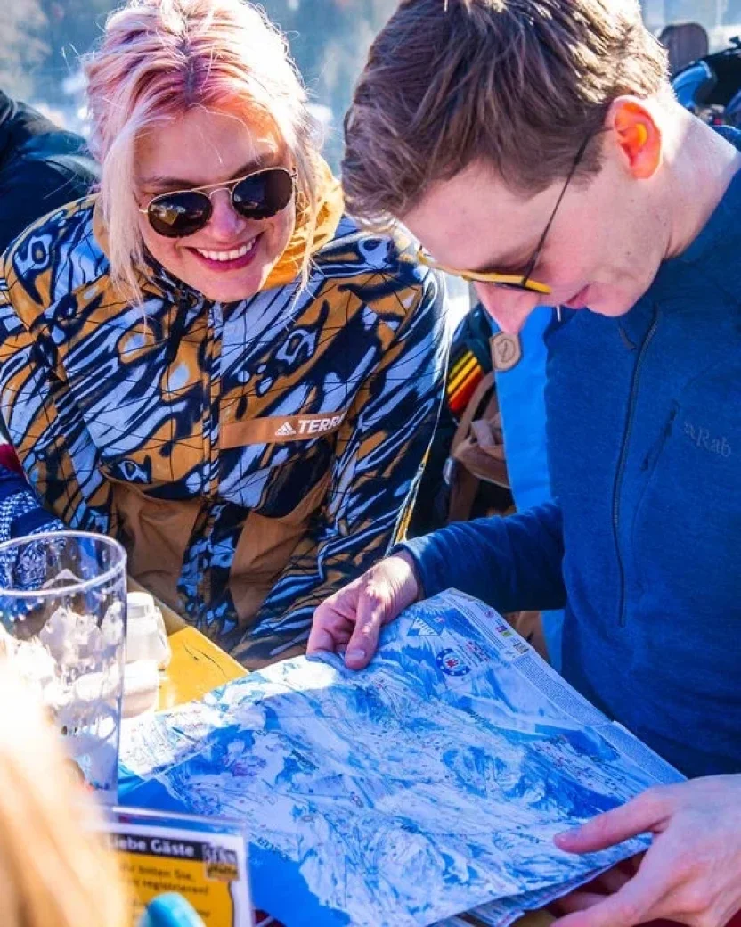 Wintersporters bekijken samen de pistekaart tijdens een pauze op een zonnig terras.