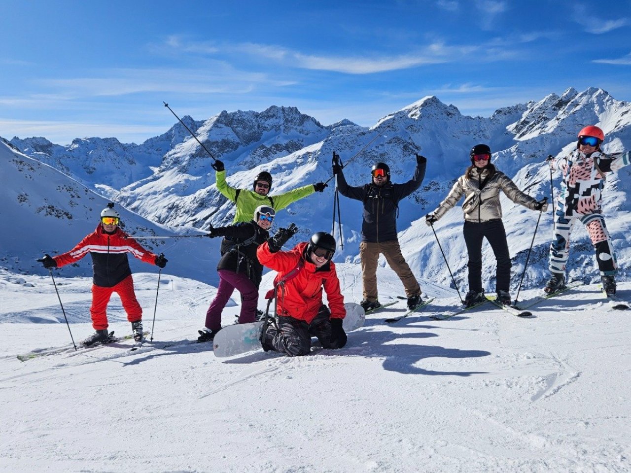 Wintersporters poseren voor de foto in de zon bovenaan de piste met besneeuwde bergen op het achtergrond.