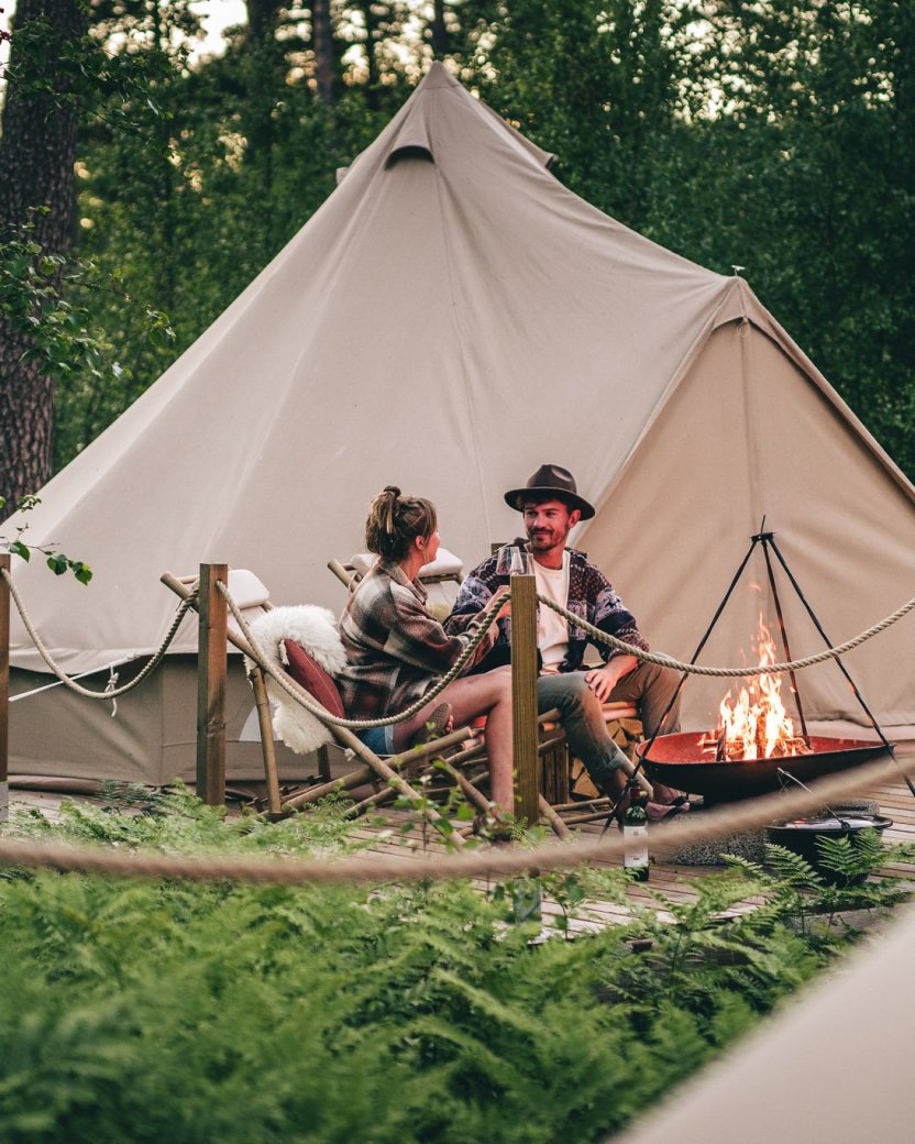 Twee reisgenoten zitten samen bij het kampvuur voor een glampingtent in het bos.