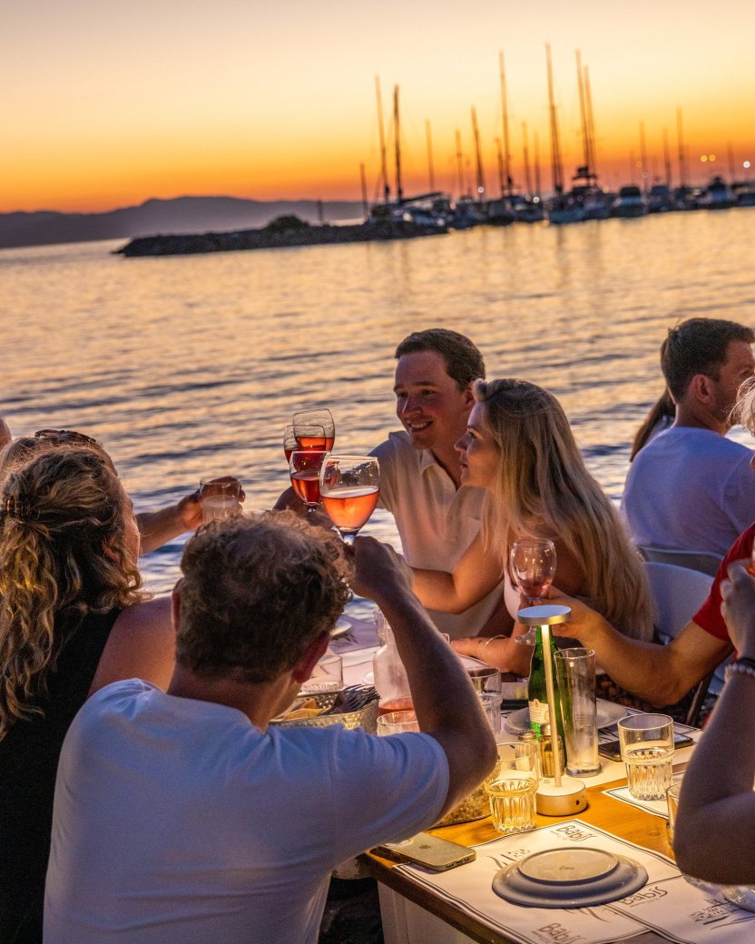 Reisgenoten proosten met een drankje tijdens het diner aan het water met zonsondergang op het achtergrond.