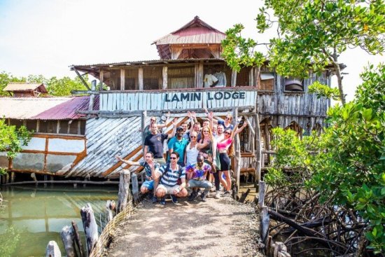 Groep reisgenoten poseert bij een houten lodge in Gambia.