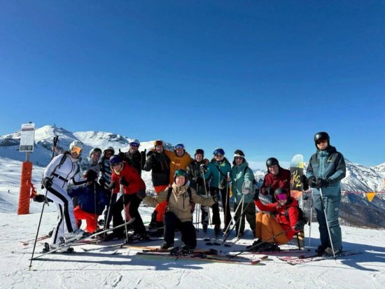 Groep wintersporters poseert bovenaan de piste met blauwe lucht en besneeuwde bergen op het achtergrond.